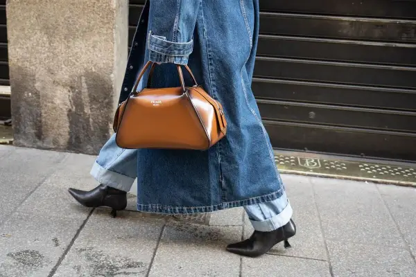 Une femme élégante marche dans la rue, portant un jean retroussé, une veste en jean et un sac à main marron. Le style "comment retrousser son jean femme" est parfait pour cette tenue.